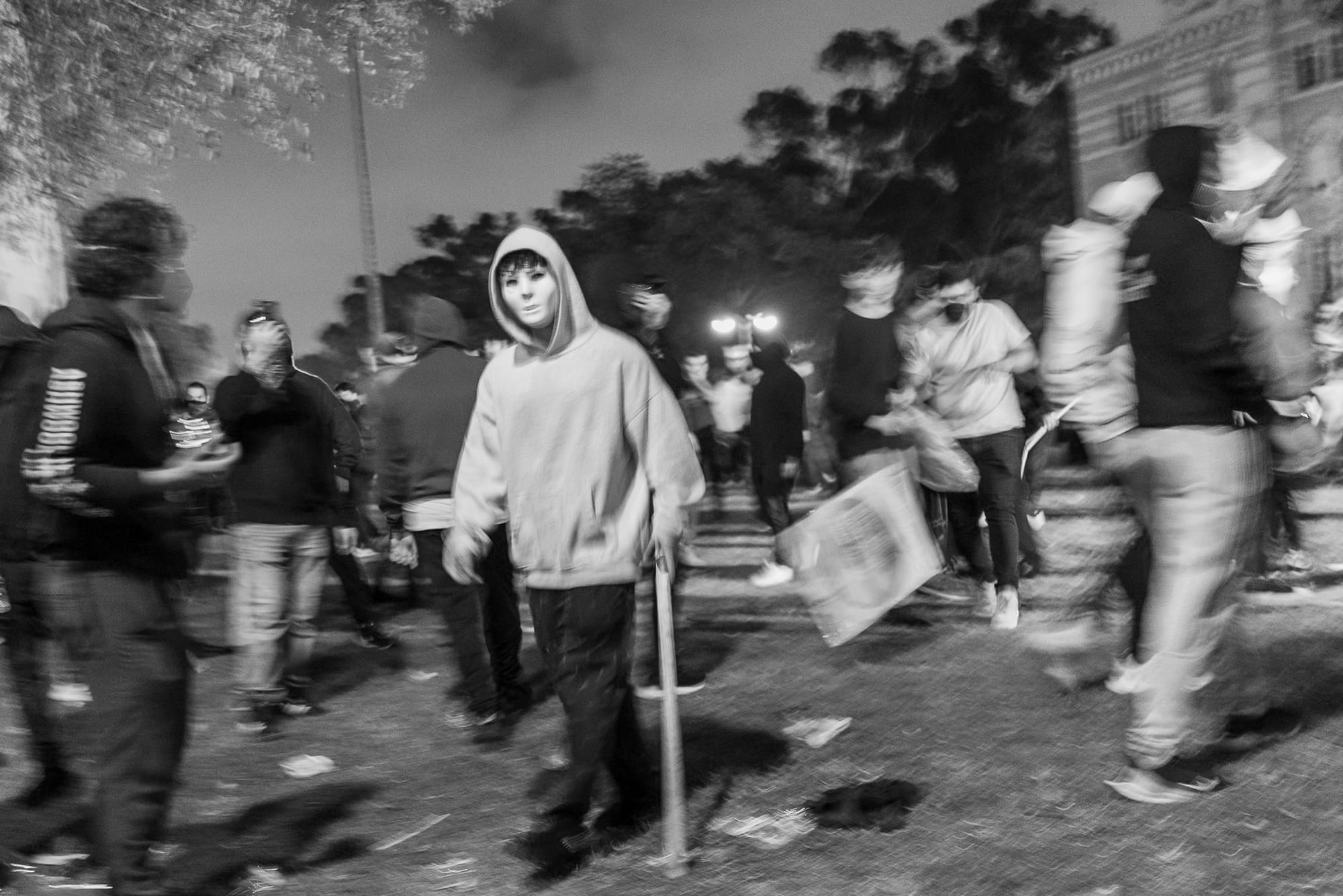 A masked person in a hoodie with the hood up hold a wooden or metal rod in his hand. People stand all around him.