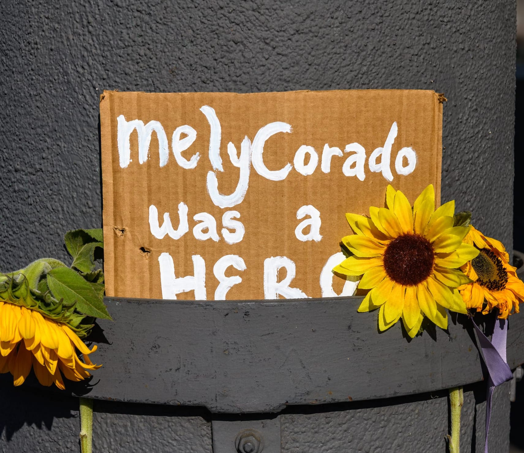 A painted cardboard sign sits on a pole outside the Silverlake Trader Joes surrounded by sunflowers. The sign reads "Mely Corado was a hero."